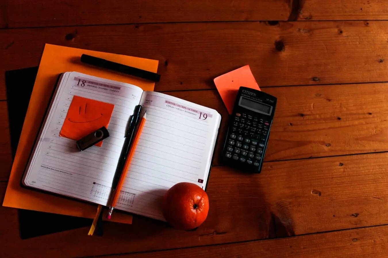 A flat-lay of an open planner, calculator, sticky notes, and an apple on a wooden desk.