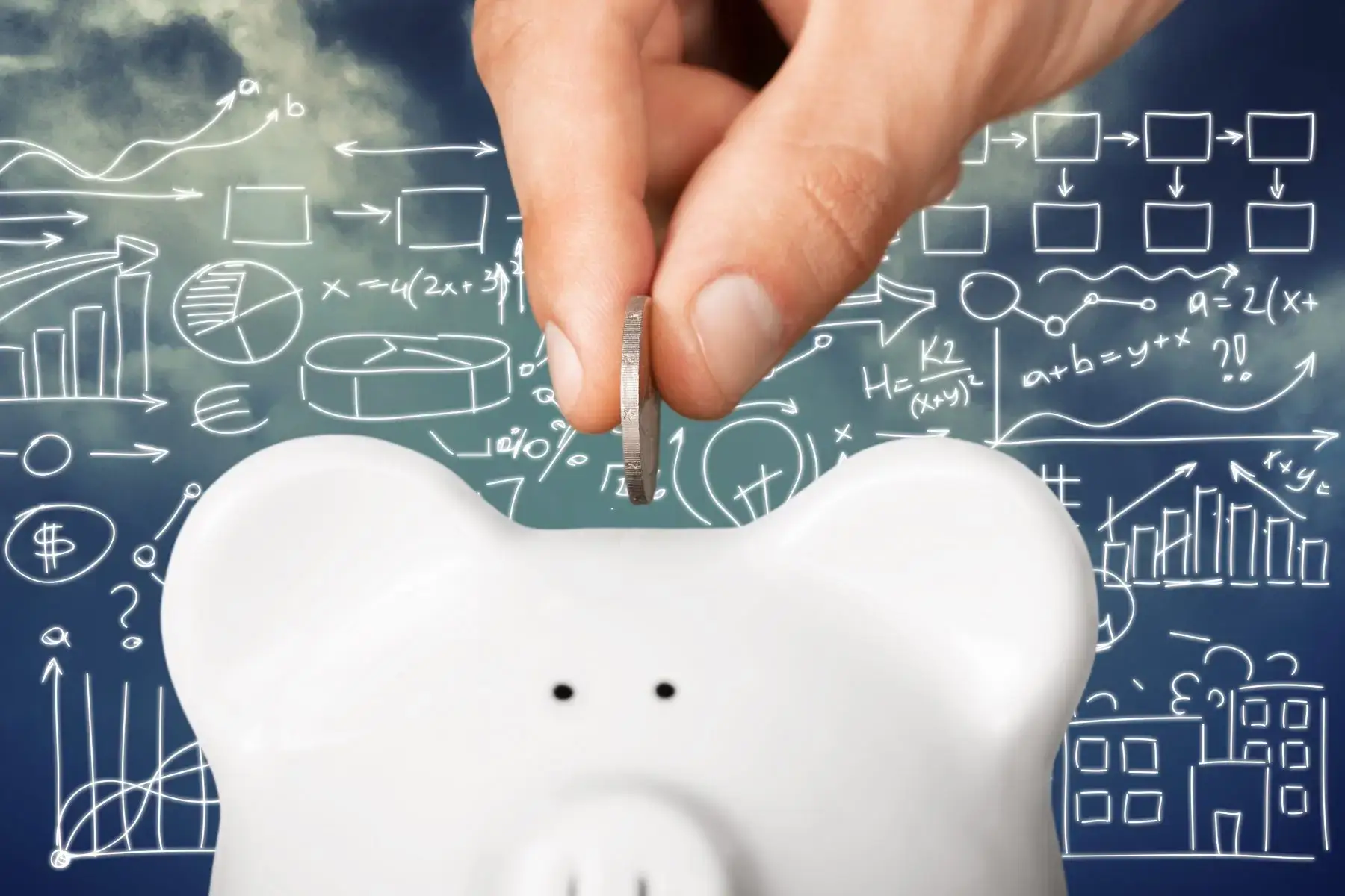 A hand dropping a coin into a piggy bank against a chalkboard of financial charts, representing savings.