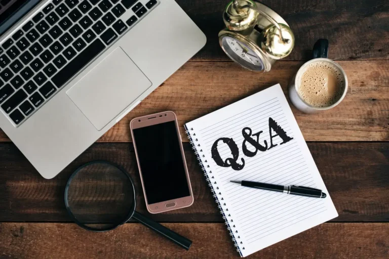A flat-lay desk with a laptop, phone, coffee, and a notebook reading Q and A.