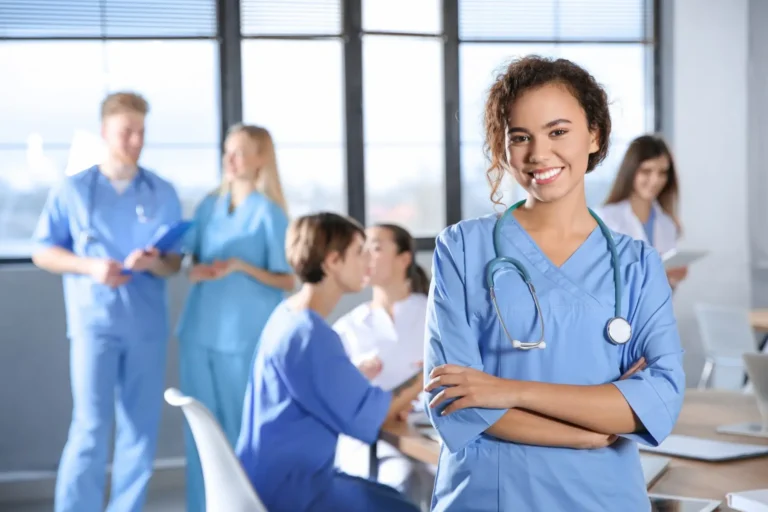 A smiling medical professional in scrubs with arms crossed, colleagues in the background.