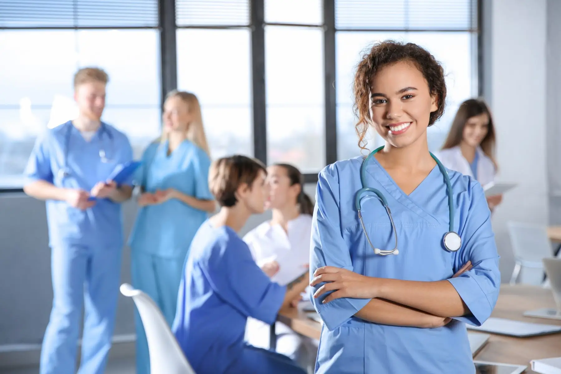 A smiling medical professional in scrubs with arms crossed, colleagues in the background.