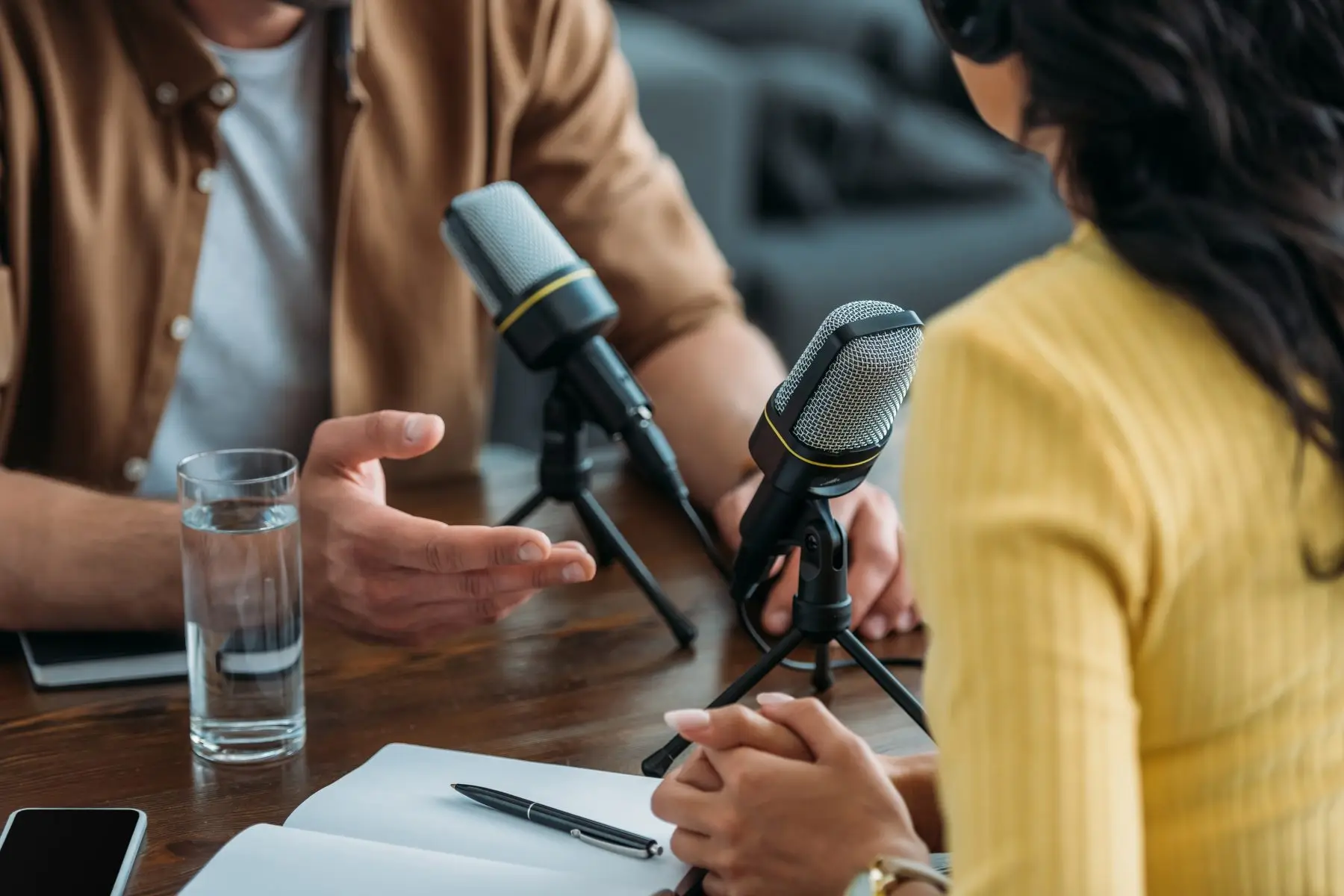 Two podcast hosts talking at a table with microphones during a recording.