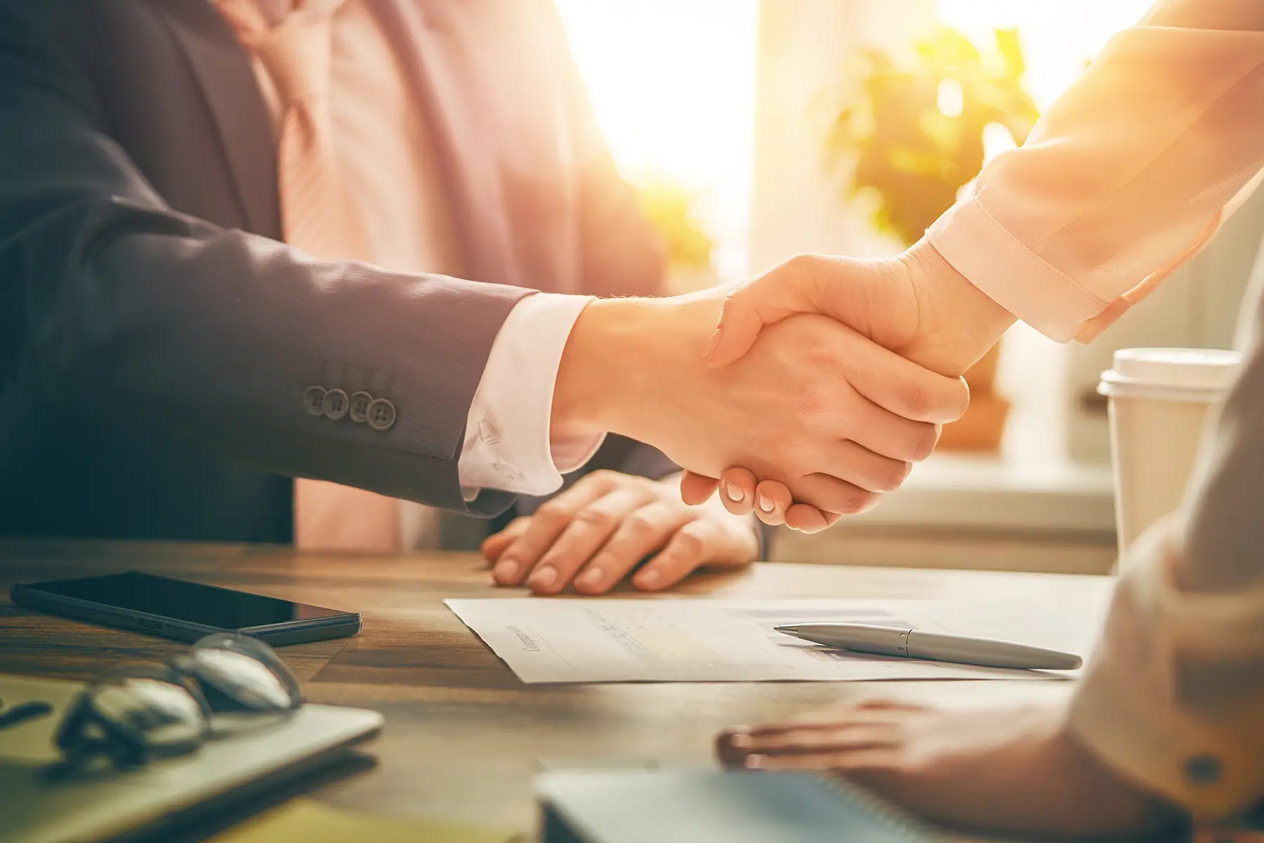 Two people shaking hands across a desk in warm sunlight.