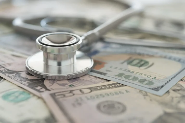 A stethoscope resting on a spread of U.S. hundred-dollar bills, representing healthcare costs.