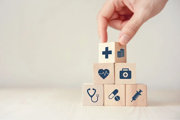A hand stacking wooden blocks printed with healthcare icons into a pyramid.