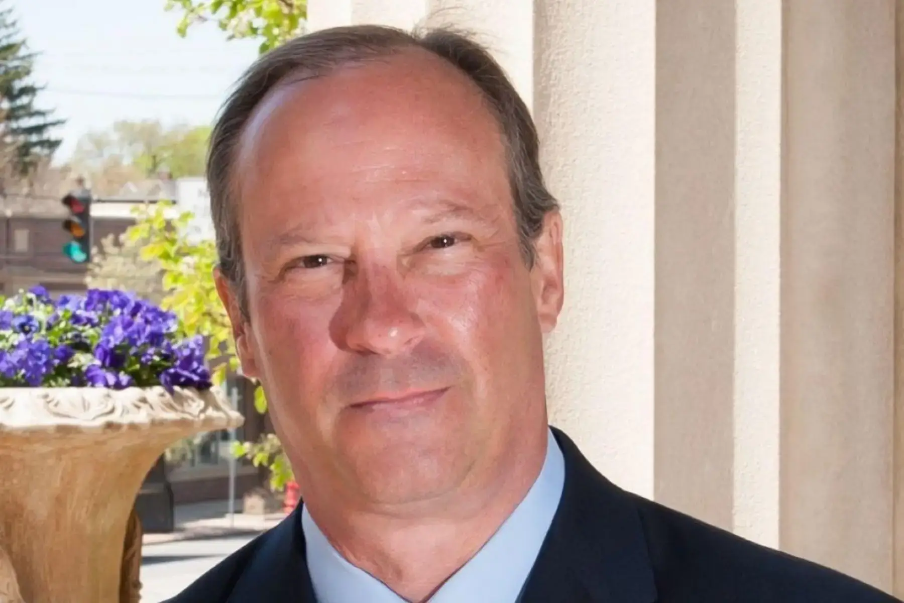A headshot of a smiling man in a dark suit outdoors.