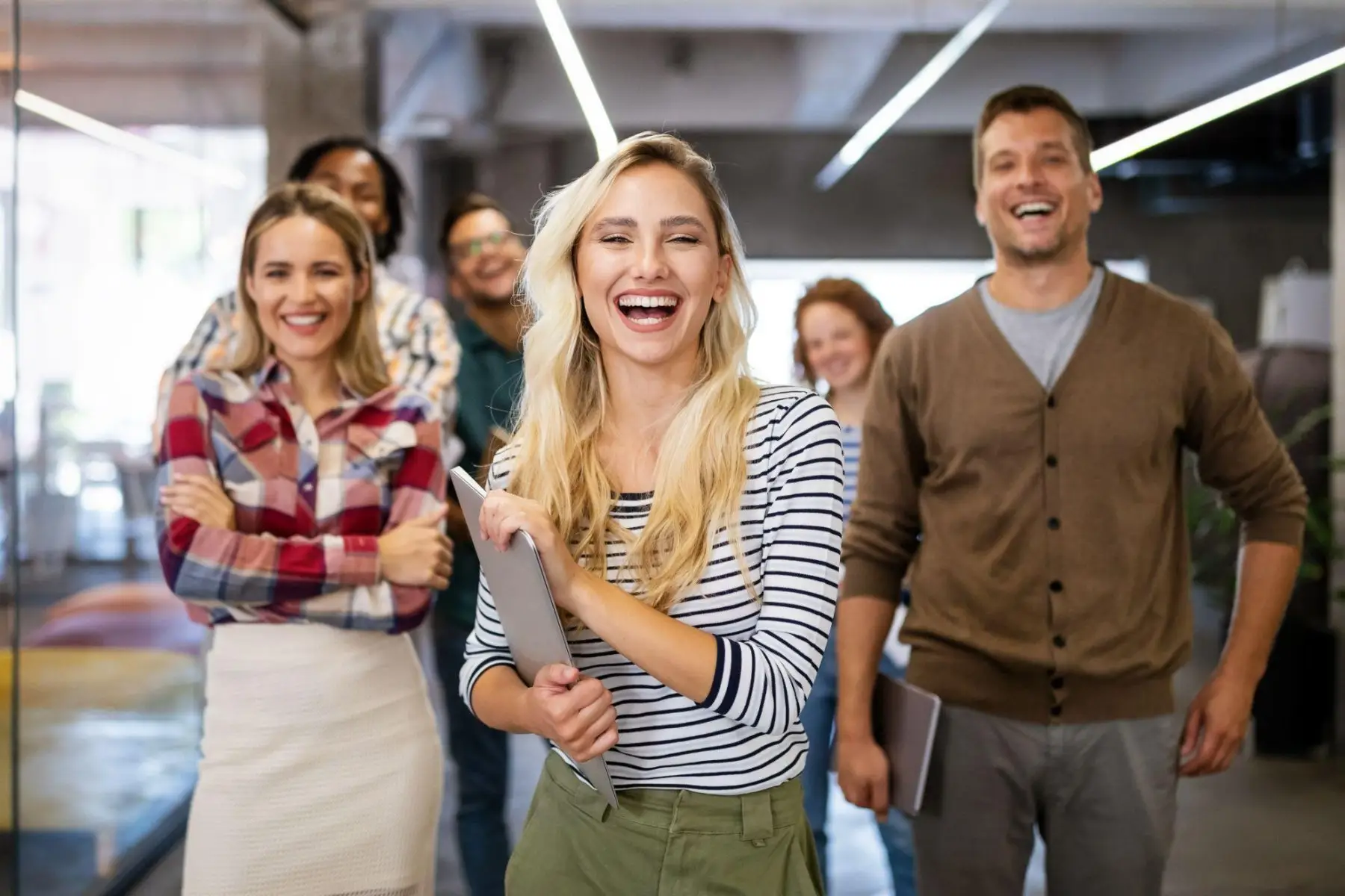 A group of cheerful colleagues walking down an office hallway, one laughing in front.