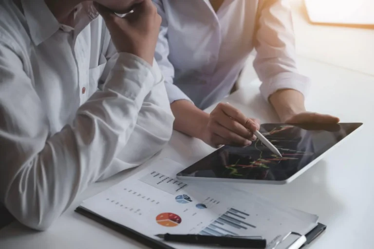 Two people reviewing financial charts on a tablet and printed reports at a desk.