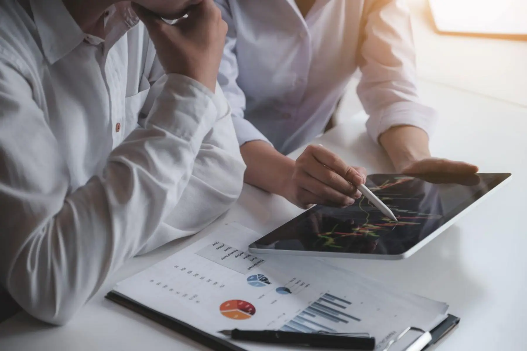 Two people reviewing financial charts on a tablet and printed reports at a desk.