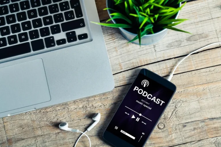 A smartphone displaying a podcast app on a desk beside a laptop and earbuds.