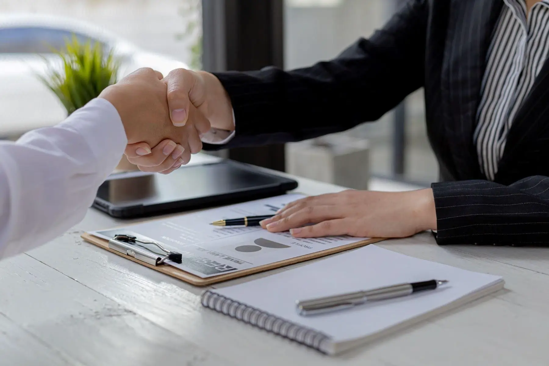 Two people shaking hands across a desk with a clipboard and pen.