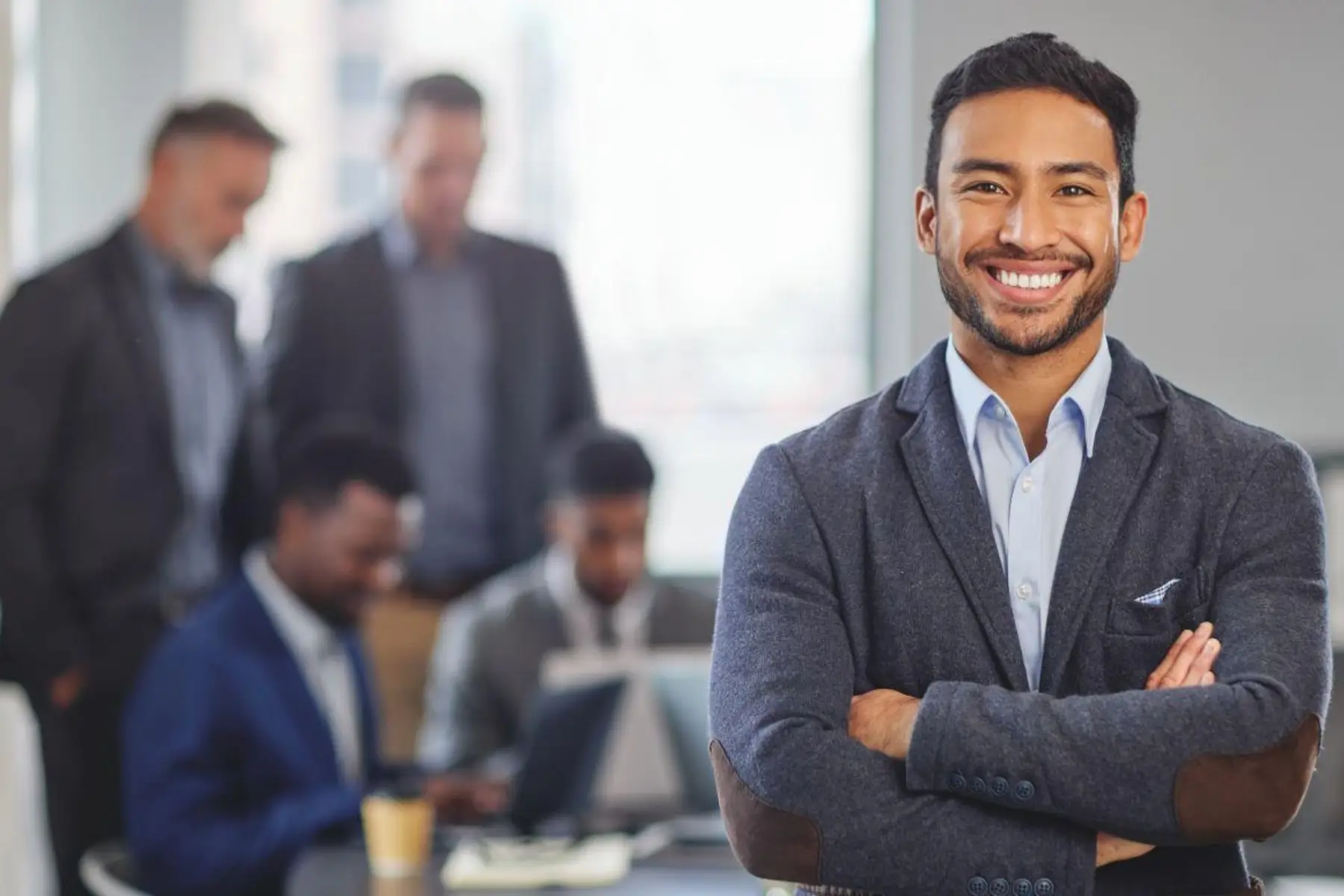 A smiling businessman with arms crossed as colleagues work in the background.