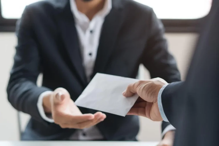 One person handing a white envelope to another across a desk.