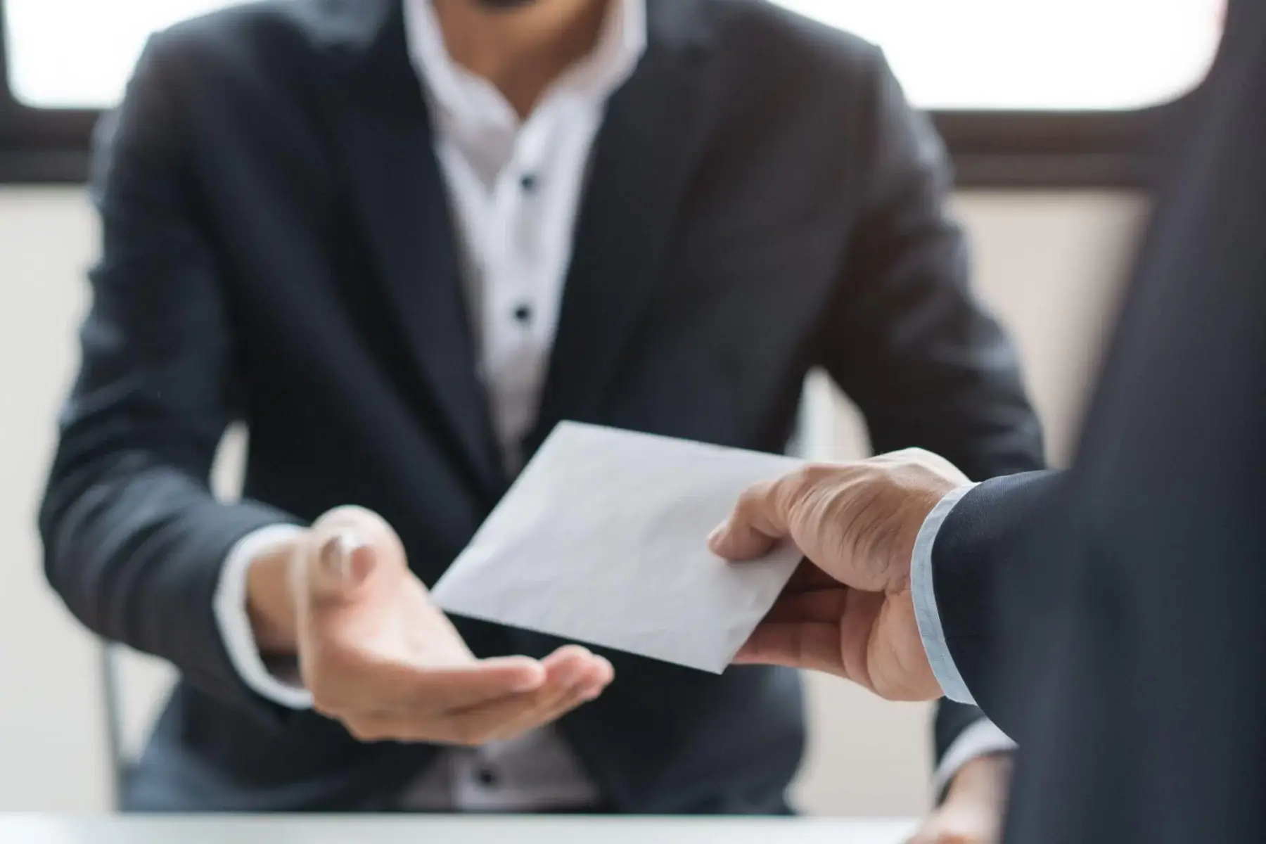 One person handing a white envelope to another across a desk.