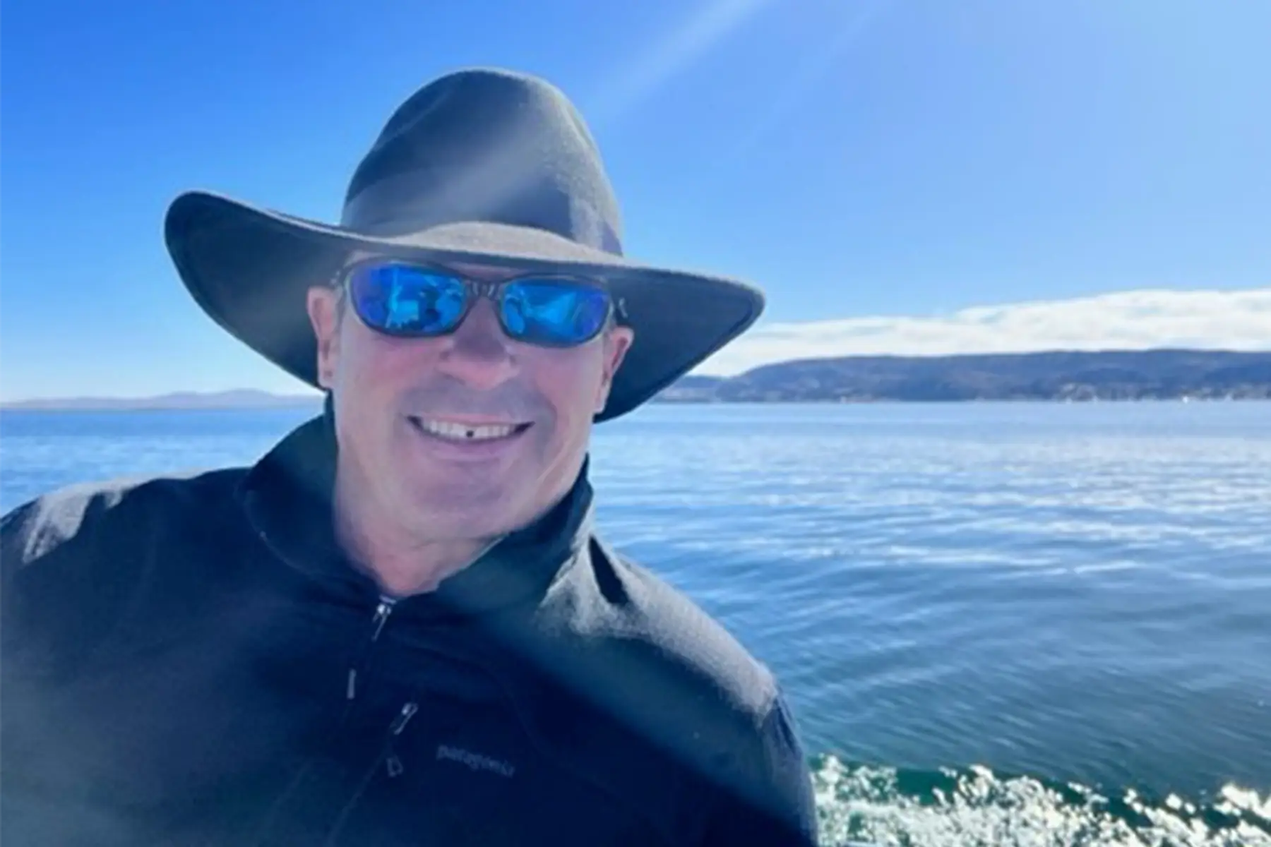 A smiling man in a hat and sunglasses on a boat, with water and hills behind him.