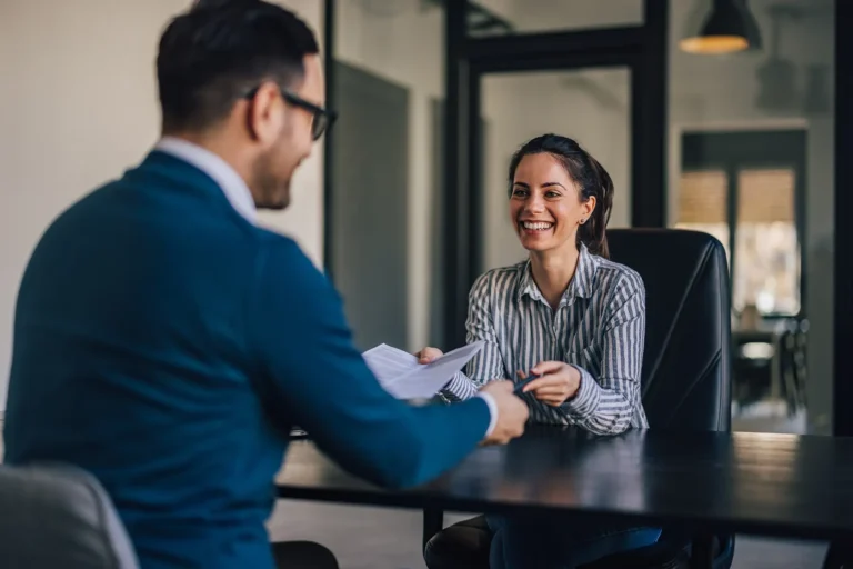 A businessman and a smiling woman talking across a desk during a meeting.