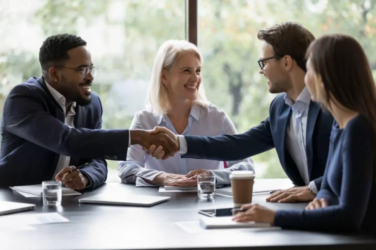 Two businessmen shaking hands across a table while colleagues look on during a meeting.