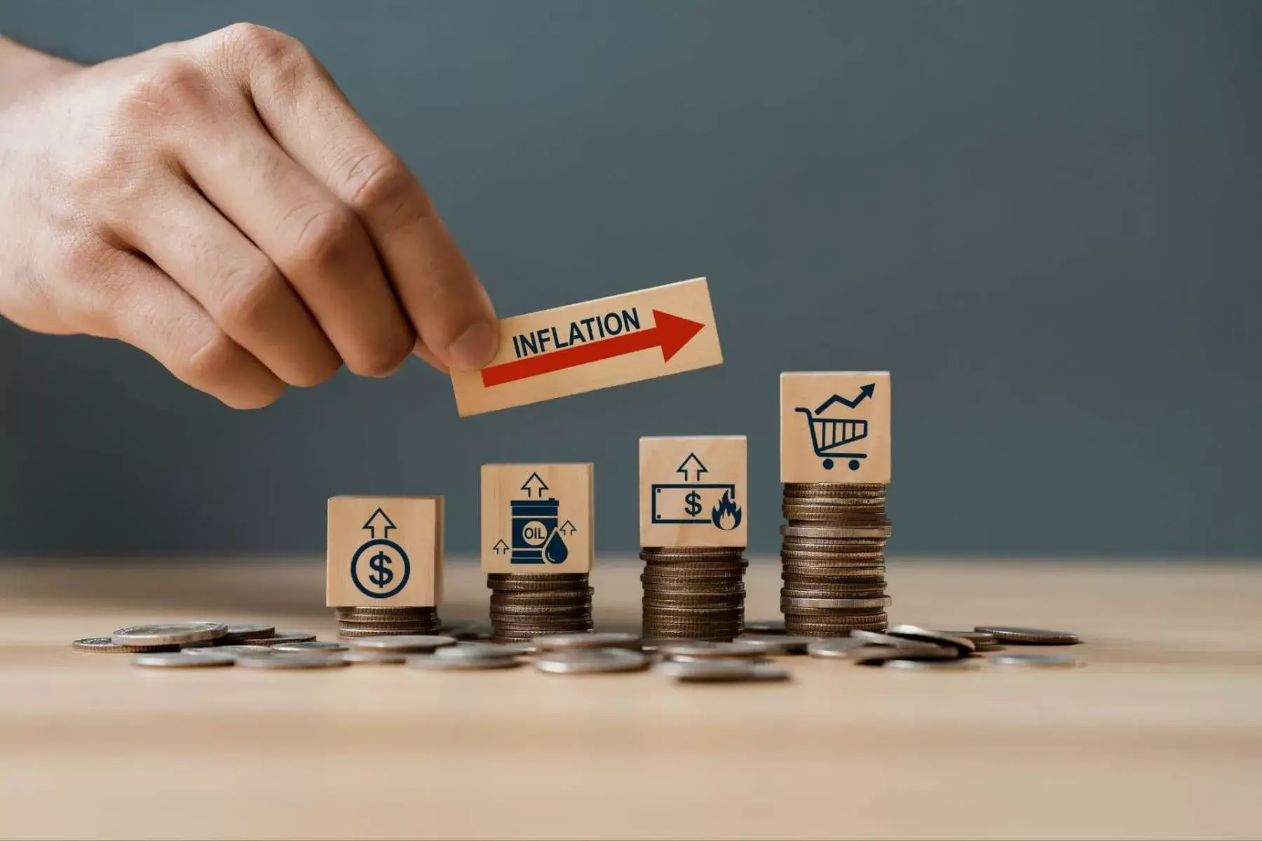 A hand placing an inflation arrow block atop rising stacks of coins with shopping icons, representing rising costs.