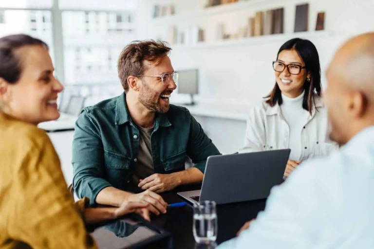 A group of colleagues smiling and chatting around a laptop in a casual meeting.