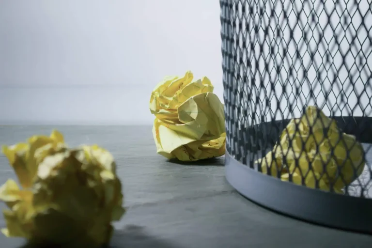 Crumpled yellow paper balls beside a mesh wastebasket, with some missing the bin.