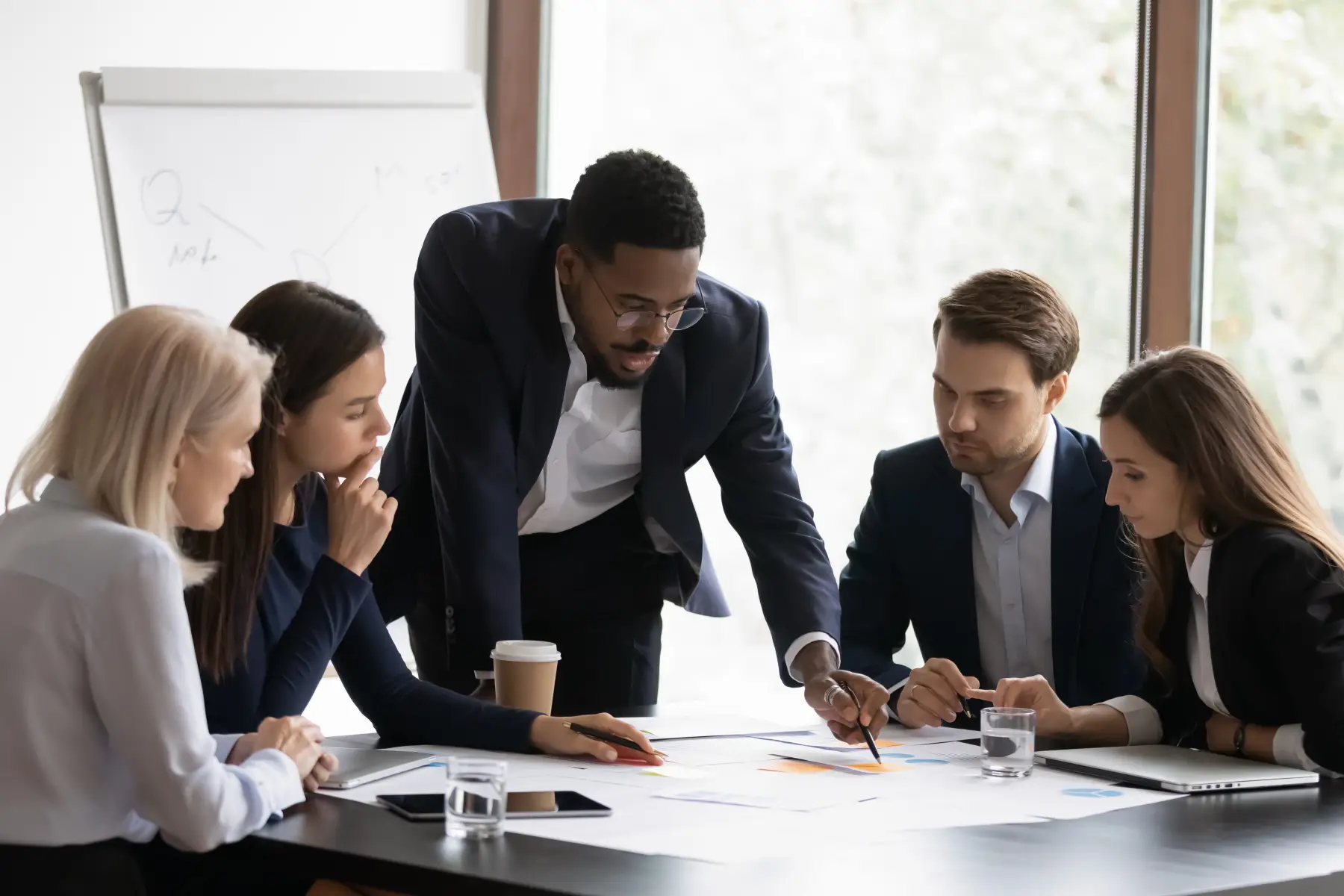 A man leaning over a conference table presenting to seated colleagues during a meeting.