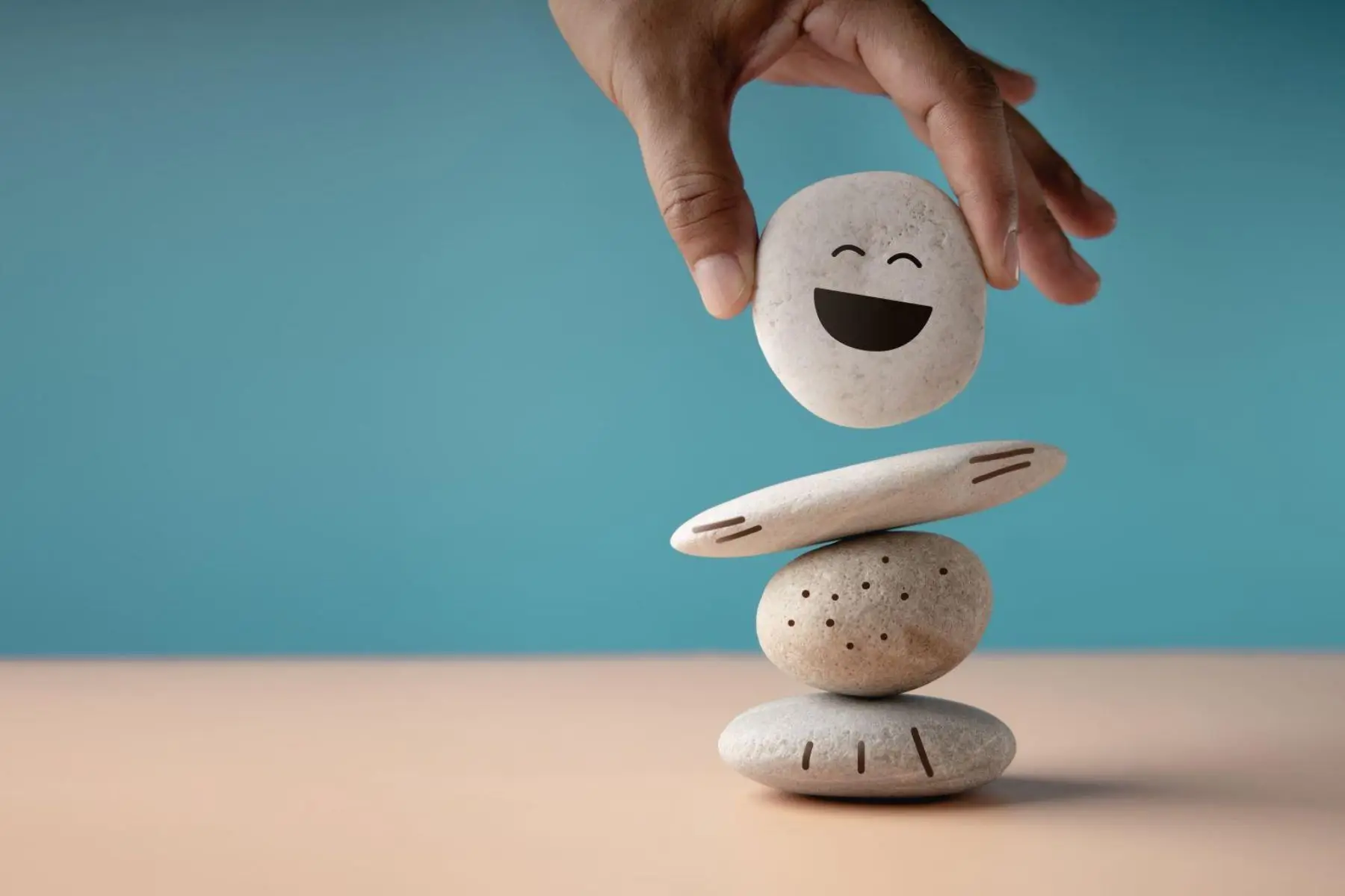 A hand placing a smiling-face stone atop a balanced stack of pebbles, symbolizing well-being and balance.