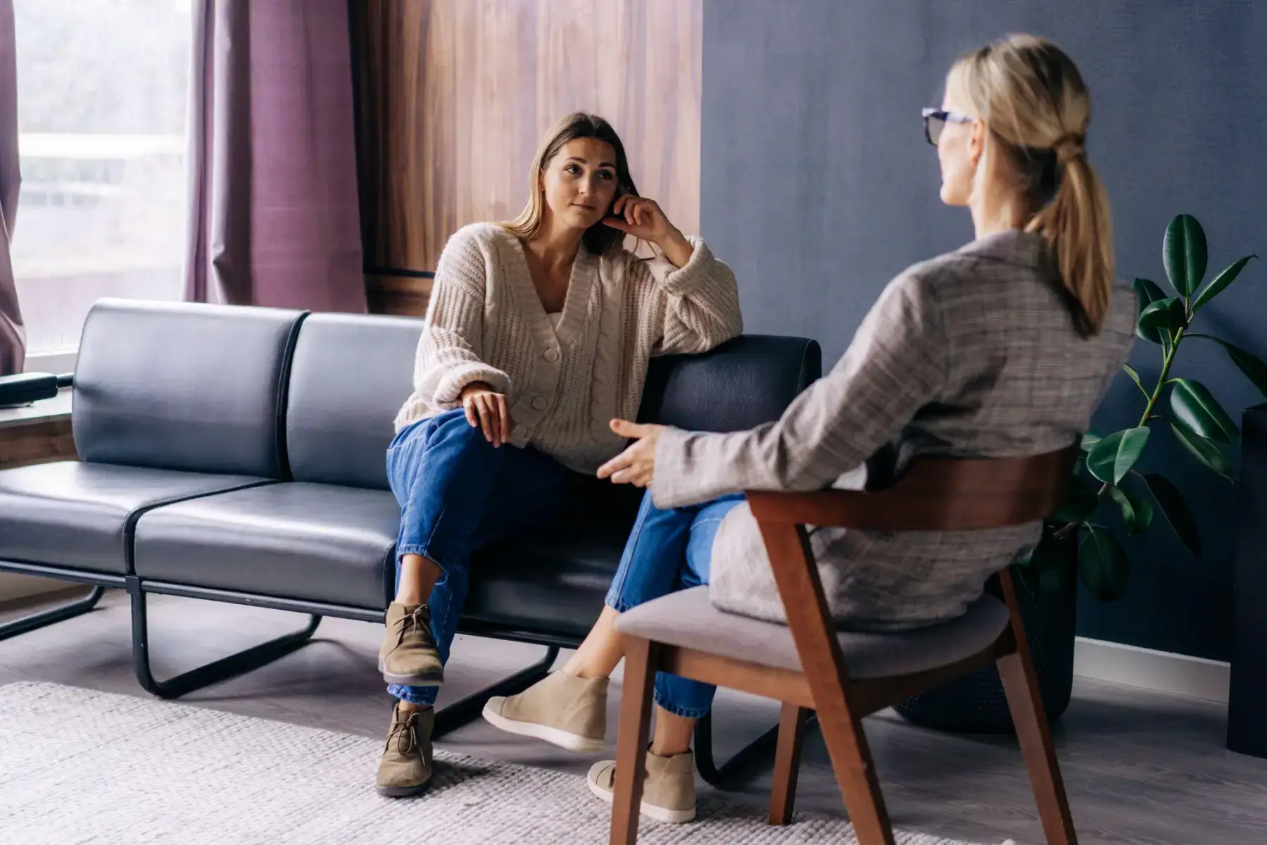 Two women in conversation in a comfortable counseling setting, one seated on a sofa.
