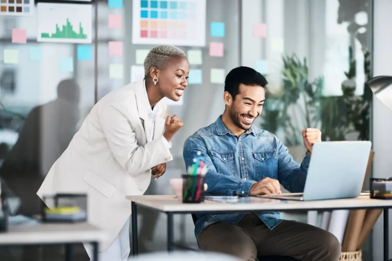 A woman and a man celebrating with raised fists at a laptop in an office.