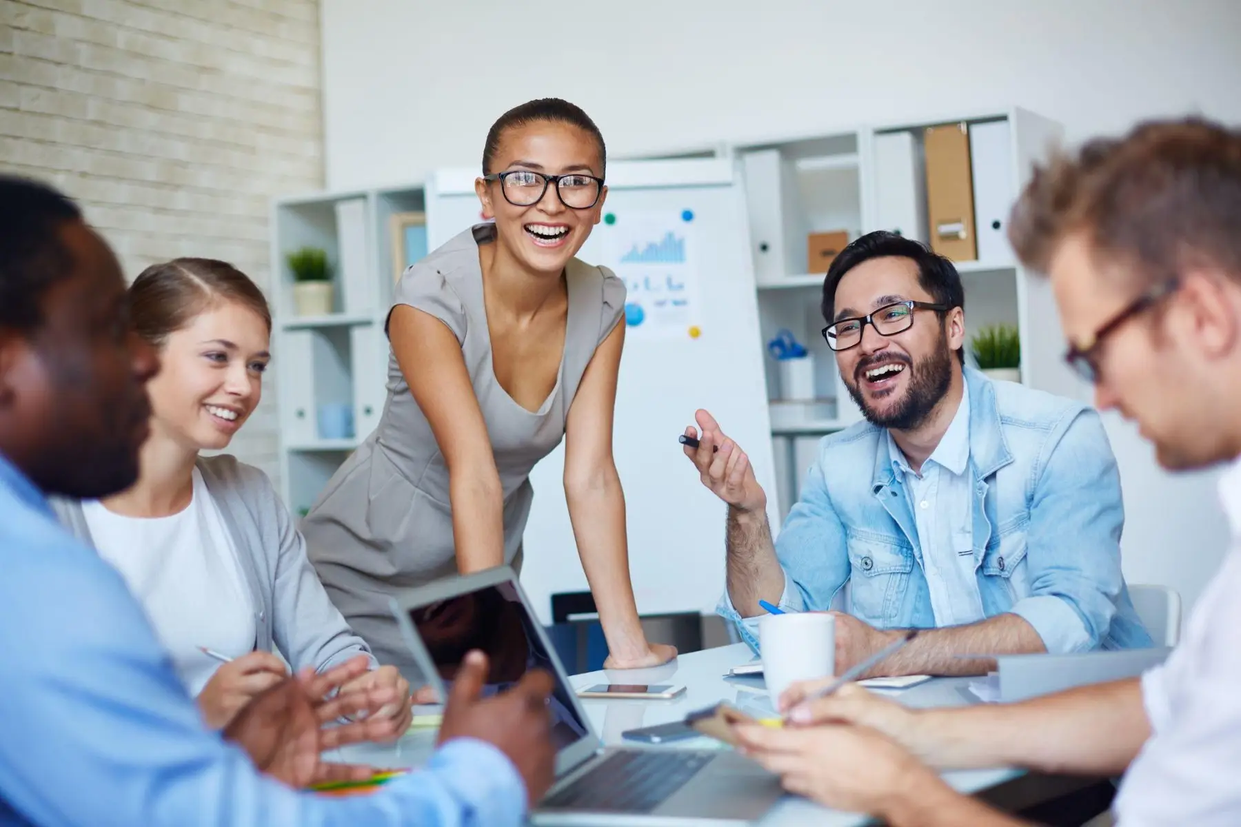 A woman presenting at a flip chart to a smiling group around a meeting table.
