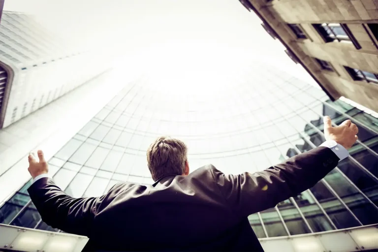 A businessman with arms outstretched looking up at towering skyscrapers.