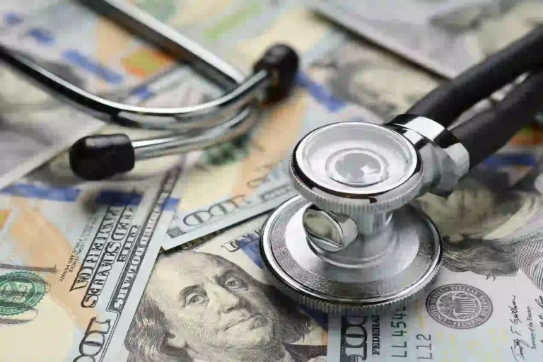 A stethoscope resting on a spread of U.S. hundred-dollar bills, representing healthcare costs.