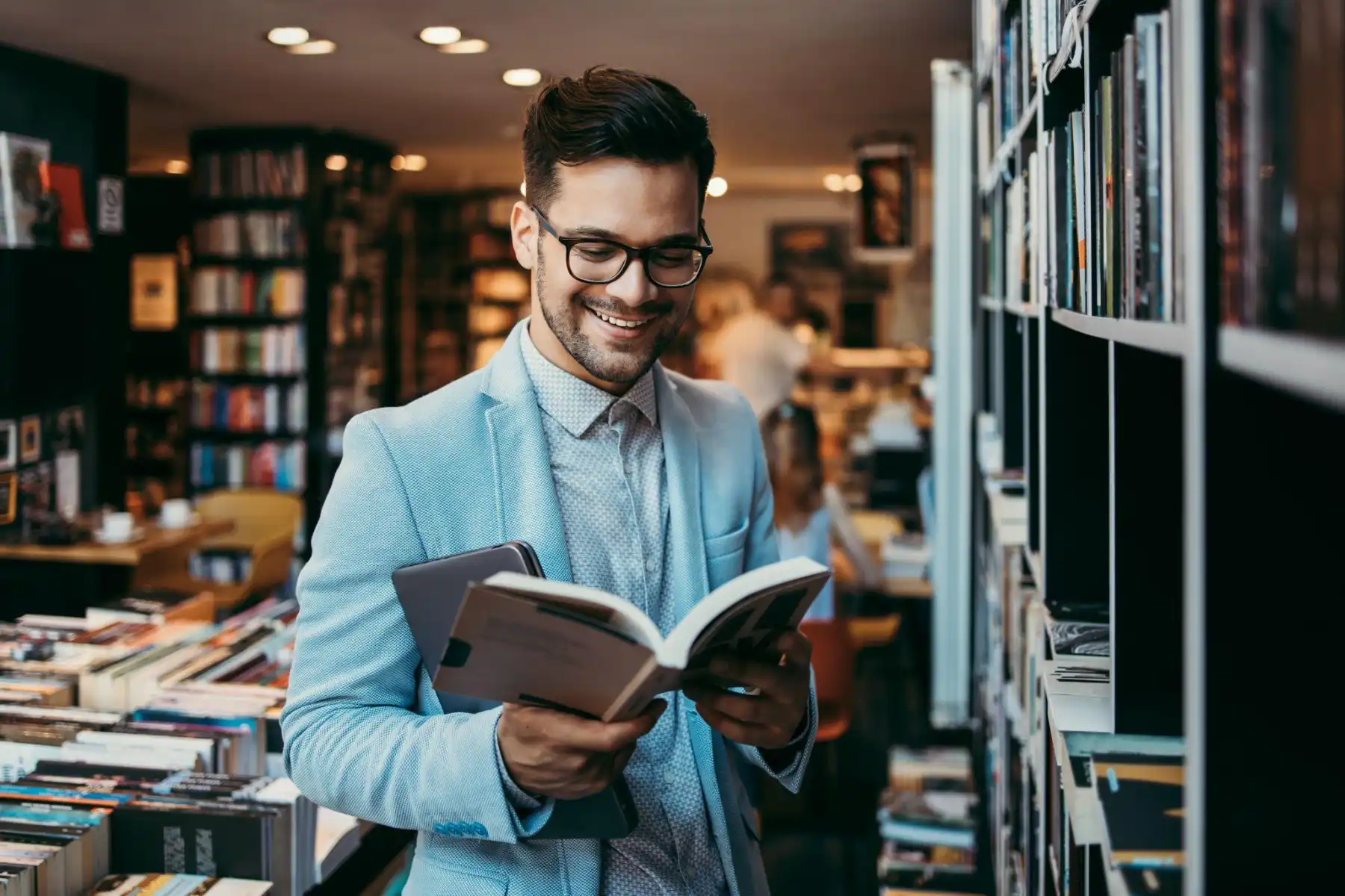 A smiling man in a blue suit reading a book in a bookstore.