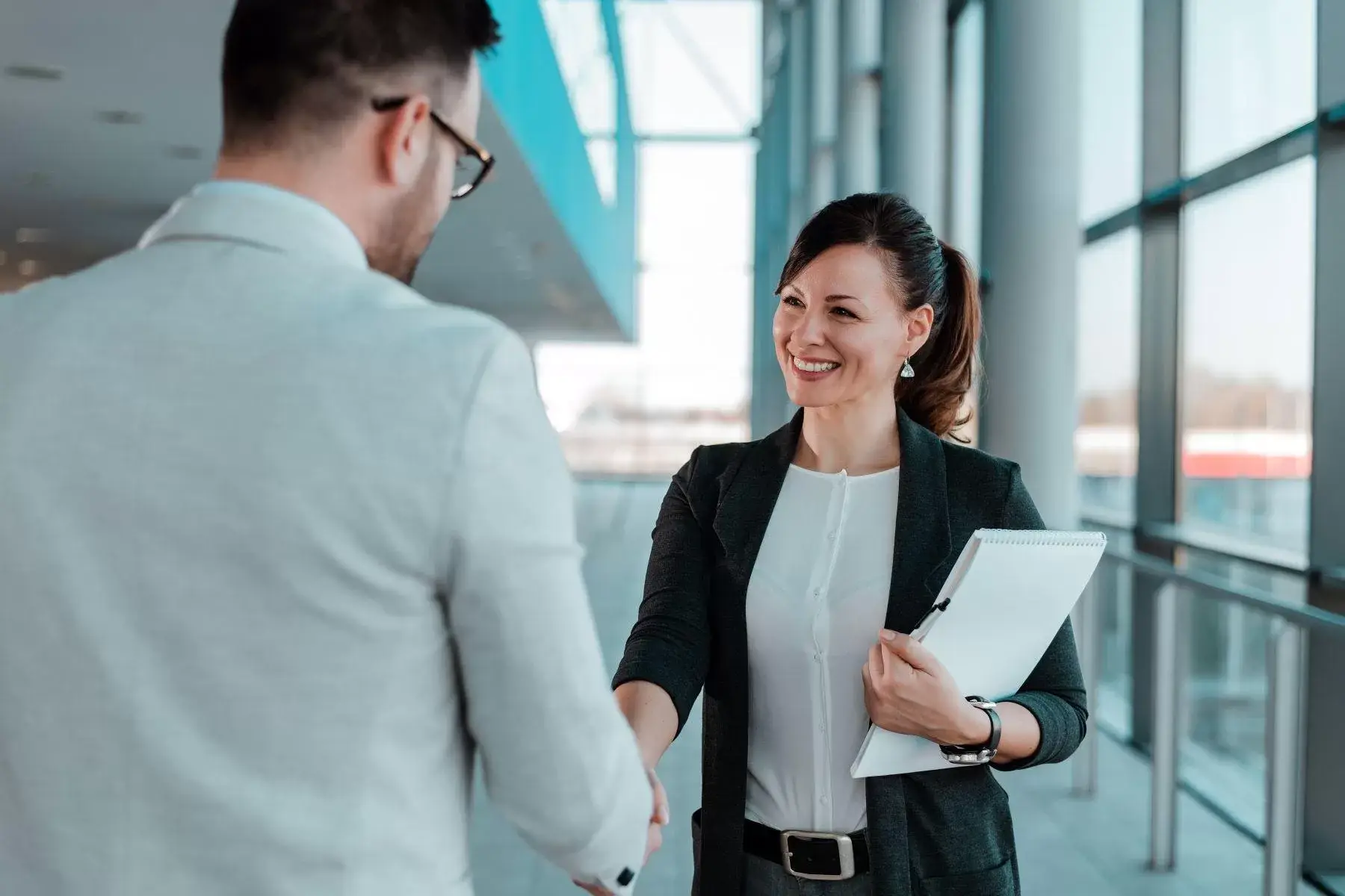 A businesswoman holding a folder shaking hands with a man in a modern lobby.