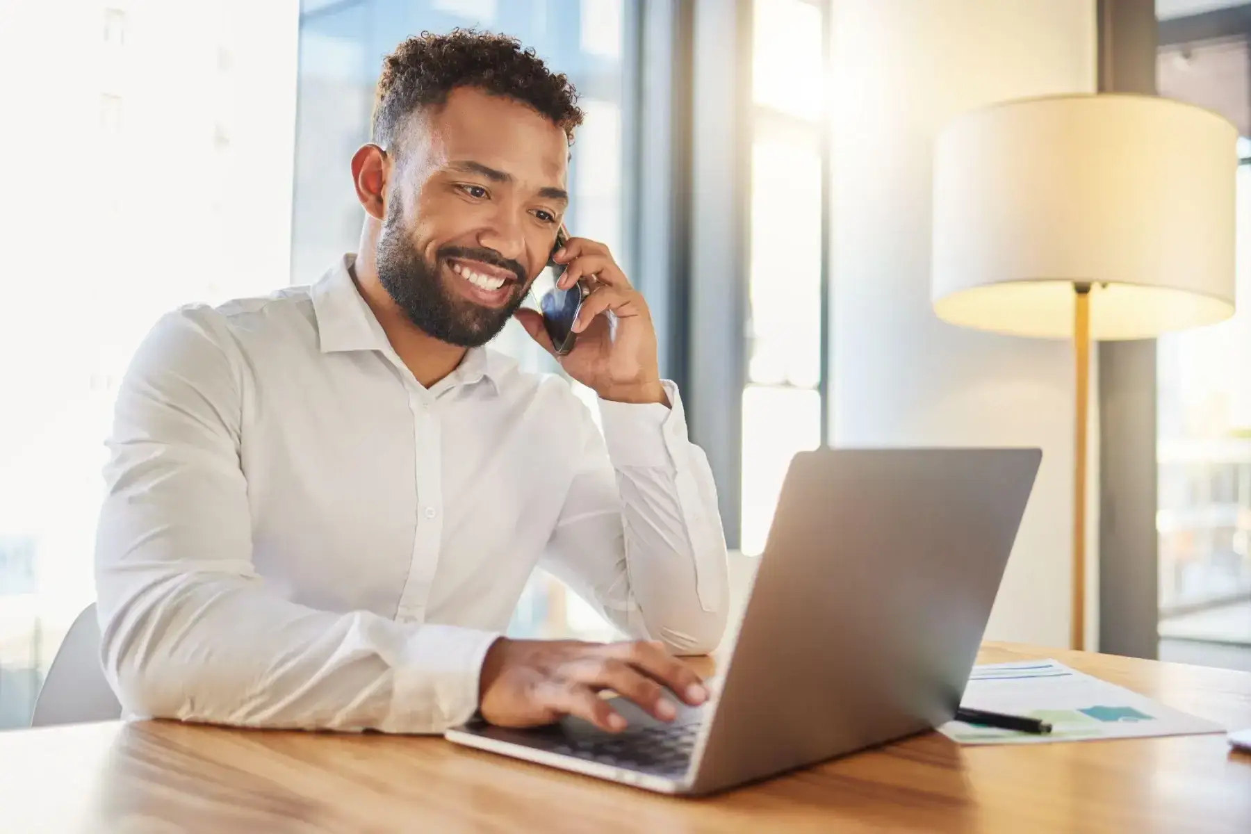 A smiling man talking on the phone while working at a laptop in an office.