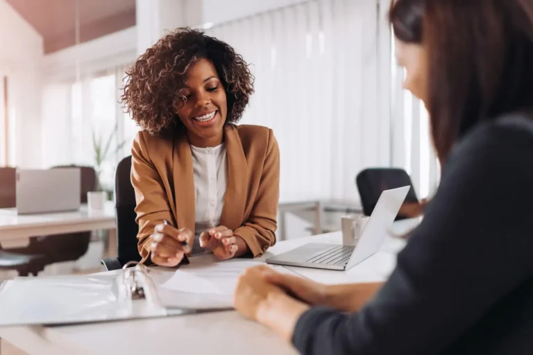 A businesswoman talking with a client across a desk in a bright office.