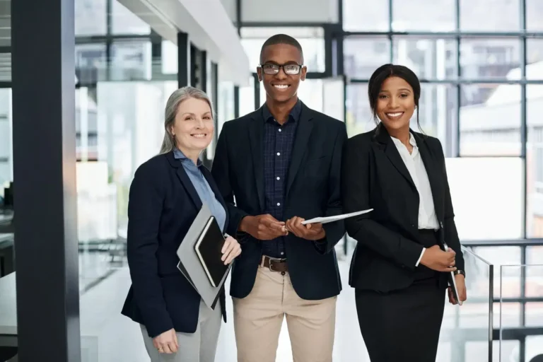 Three business professionals standing together holding folders and tablets, smiling.