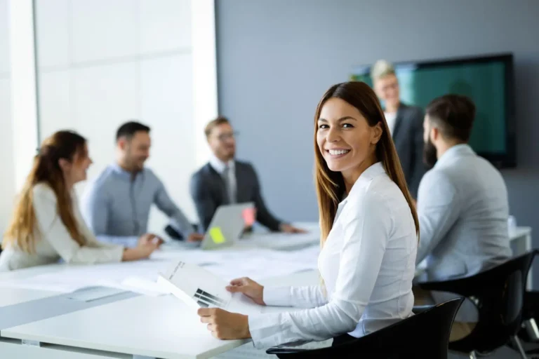 A smiling businesswoman at a conference table, with colleagues meeting in the background.