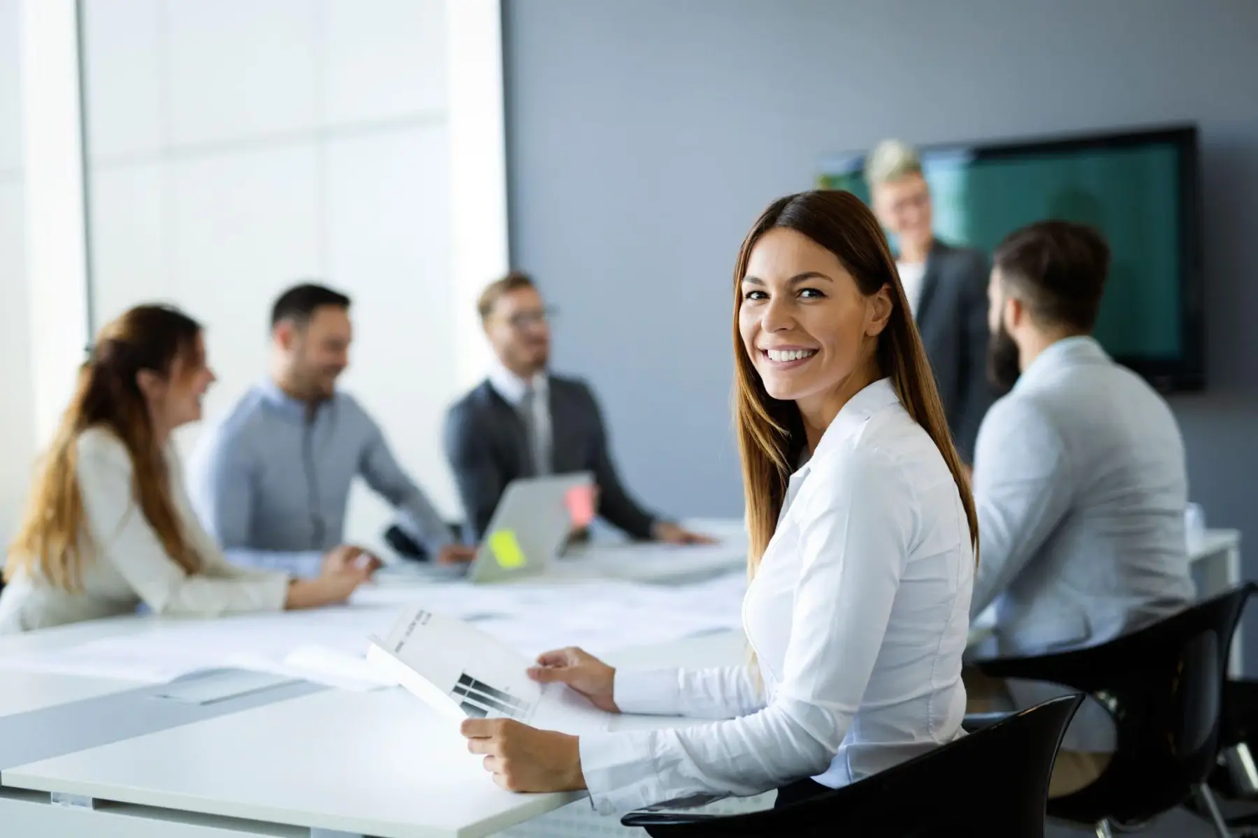 A smiling businesswoman at a conference table, with colleagues meeting in the background.