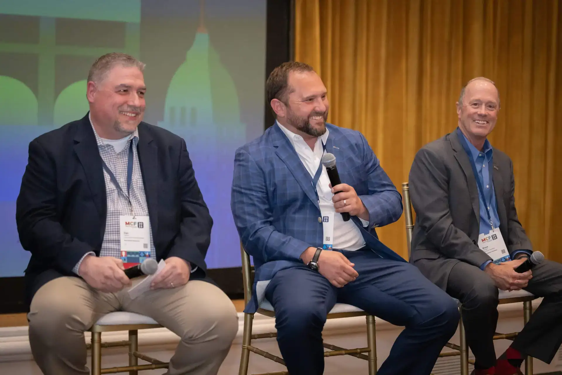 Three panelists seated on stage at a conference, one speaking into a microphone.