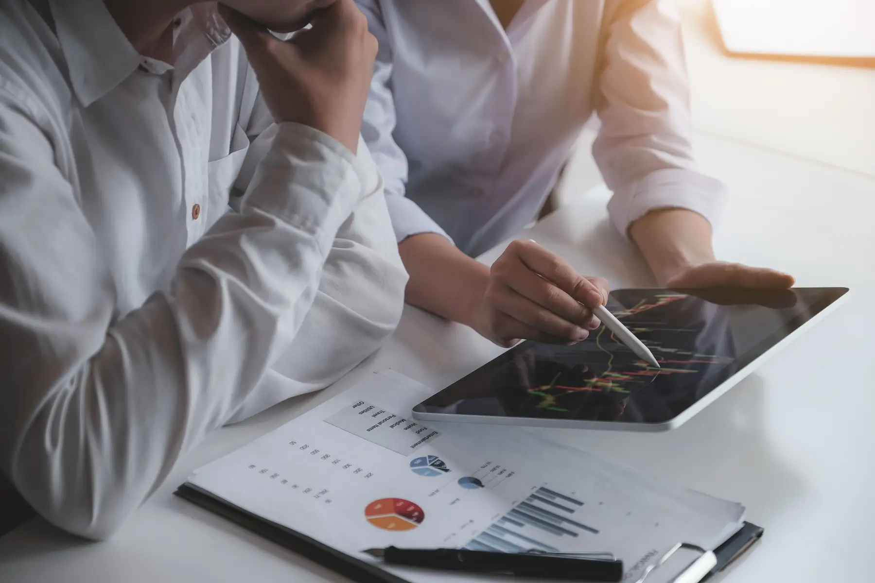 Two professionals reviewing financial charts and data on a tablet and paper reports at a desk.