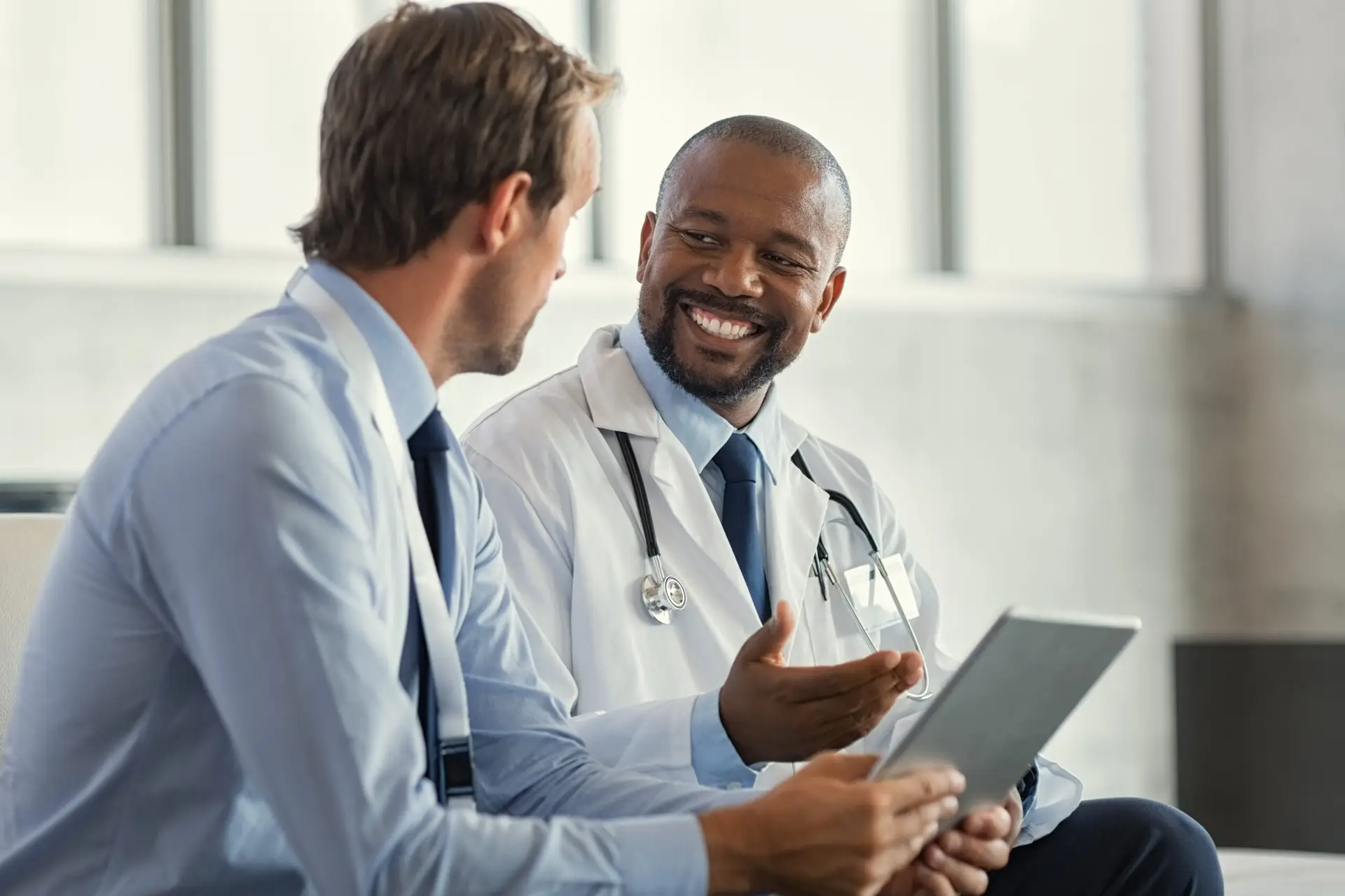 A smiling doctor holding a tablet talking with a male patient.
