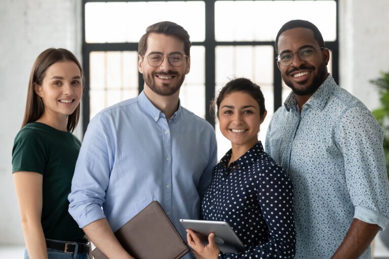 Four smiling professionals standing together in an office, one holding a tablet.
