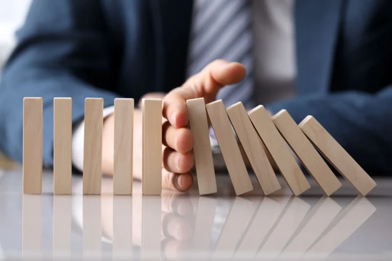 A hand stopping a row of falling wooden dominoes, symbolizing risk prevention.