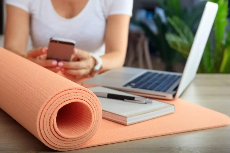 A woman with a rolled yoga mat at her desk, checking her phone beside a laptop.