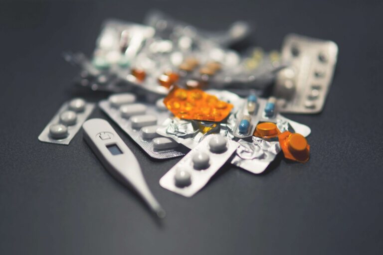 A pile of assorted blister-pack medications and capsules with a thermometer.
