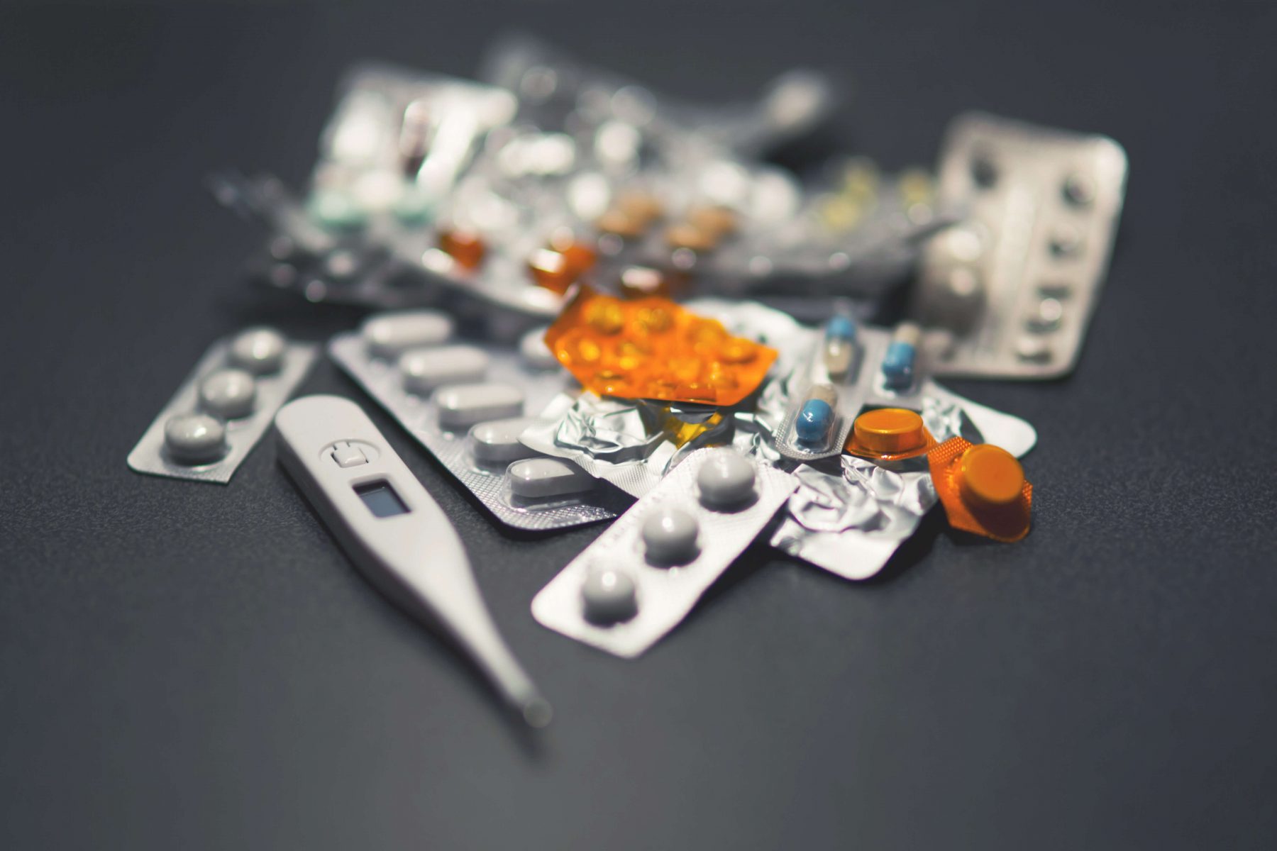 A pile of assorted blister-pack medications and capsules with a thermometer.