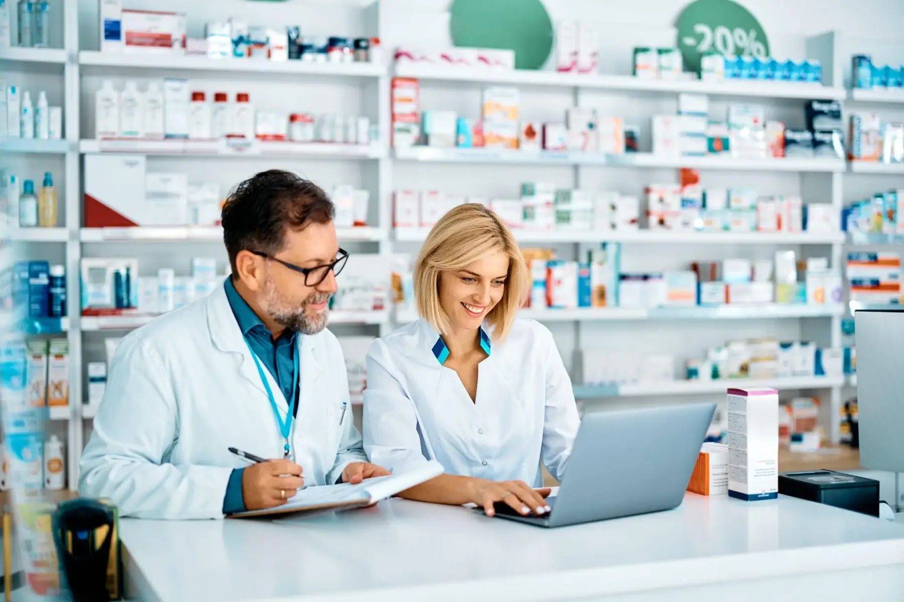 Two pharmacists in white coats reviewing information on a laptop behind a pharmacy counter.