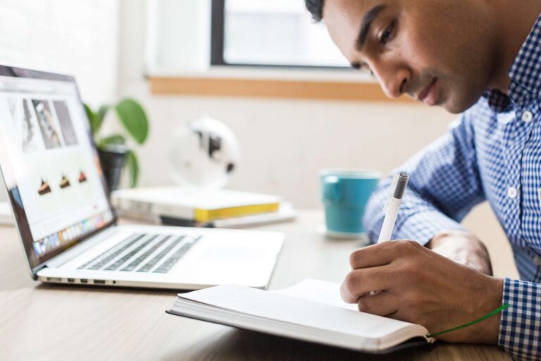 A young man writing in a notebook beside a laptop at a desk.