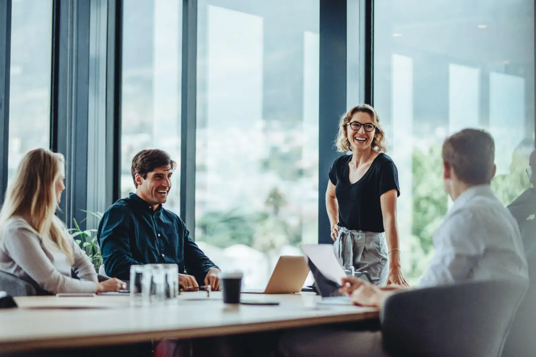 A woman leading a meeting with colleagues around a table by large office windows.